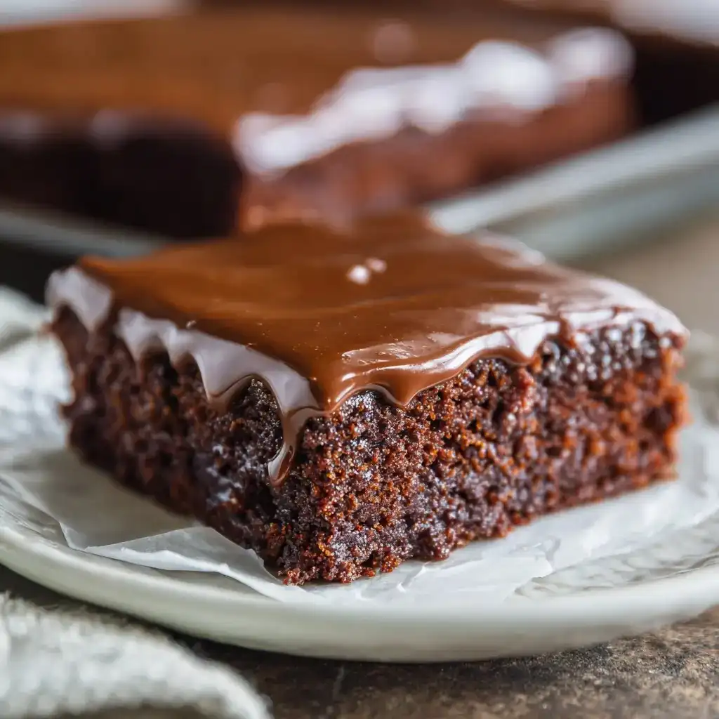 Slice of Texas Sheet Cake with warm chocolate frosting on a white plate.