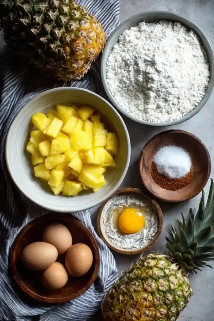 Overhead view of fresh pineapple chunks, flour, eggs, sugar, and brown sugar arranged for making pineapple upside-down cake.